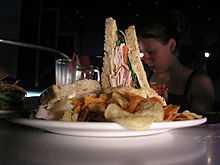 A photograph of a triangular sandwich with tomato, lettuce, ham, and cheese beside a pile of potato chips on a white plate all in front of a girl with her hair in a bun