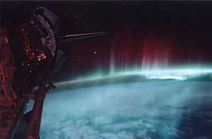 The lower half is the blue-white planet in low illumination. Nebulous red streamers climb upward from the limb of the disk toward the black sky. The Space Shuttle is visible along the left edge.