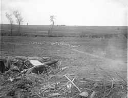 Two soldiers operate a field gun from a dug-in position in an open field and reinforced with sandbags. Entrenching tools, spoil and broken vegetation lie around the emplacement, while a number of defoiliated trees stand in the middle distance.