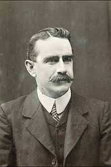 a formal black and white portrait of a male with a moustache wearing a suit