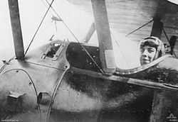 Close-up photograph of the side of an aeroplane. A man wearing a cap and with goggles on his forehead is seated in the cockpit.