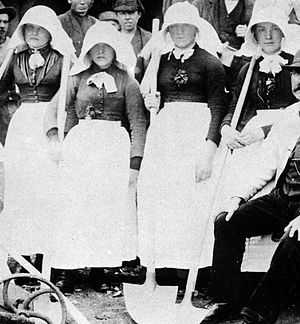 Four women wearing dark heavy clothing, bright white aprons, and long white bonnets entirely covering the sides of their heads and protruding forwards over their faces