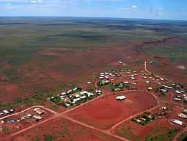 an aerial view of a small settlement, showing about seventy rooftops, red dirt roads and a dirt oval, with dry scrubland receding into the distance