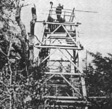 A black and white photograph of a wooden tower in the woods. A man is standing on its open deck