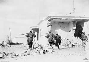 Soldiers wearing greatcoats and steel helmets with fixed bayonets run past whitewashed buildings damaged by shellfire
