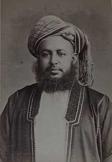 A black-and-white photograph of a man with a dark beard wearing a turban, a dark jacket, a shirt, and a belt, sitting in a chair, and looking at the viewer