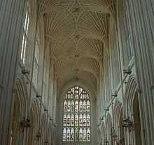 Interior of large building with a stained glass window at the far end. Above is a fan vault ceiling and on either side rows of arches.