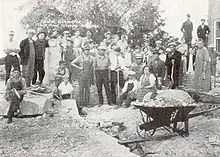 A wheelbarrow full of debris sits in the foreground, while townspeople and workers look at the unknown photographer, apparently taking a break from cleaning up debris after the bombing at the school