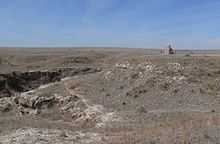 Dry grassland with a small rocky canyon; pyramidal stone monument on hill nearby