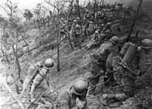 Two soldiers armed with a flame thrower are walking to the right with two soldiers armed with rifles. In the background a group of soldiers are resting over a desolate landscape