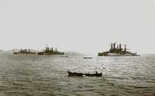 A black and white shot of three large ships near a hill-like background, with several small skiffs in the foreground.