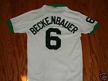 A white soccer jersey, viewed from the back; the cuffs and collar are green and on the back it is marked with the number 6 and the name "Beckenbauer".