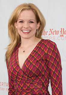 A smiling blonde woman wearing a red wrap dress, standing in front a wall with text on it