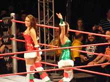 Two dark-haired Caucasian female identical twins are standing in a wrestling ring with red ropes, facing in opposite directions. They are both wearing dresses in the fashion of 'Santa Claus', although one is red and one is green.