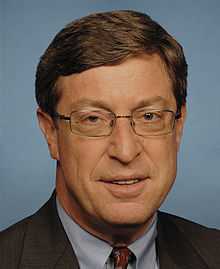 A man with brown hair and glasses wearing a black jacket, gray shirt, and red patterned tie