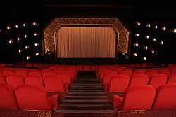 interior shot of red velvet seats facing a gilded decorated proscenium arch flanked by decorative wall lights