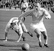 A black-and-white action shot taken in the midst of an association football match. To the viewer's right a player in light shirt, white shorts and white socks turns to his right, his eyes pointed down towards the ball. A shield bearing the letters "MFF" is prominently displayed on his shirt. Behind him, an opposing player in a white shirt can be seen stumbling mid-chase, throwing his arms forward to break his fall.