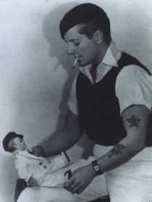 Three-quarter length portrait of a tattooed, short-haired woman in casual, male clothing. She is smoking and looking down at a doll she is holding.