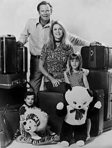 A black-and-white photograph of a man, a woman, a girl, and a boy, all looking at the viewer while surrounded by a large number of suitcases and two stuffed toys