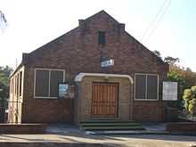 A photograph of a brown-bricked building with two wooden doors at the top of three green steps under a black number "18" all under a blue sky