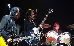 Prominent in the foreground, two guitarists concentrate on their playing, while the drummer, a little behind them to their left, toils away.