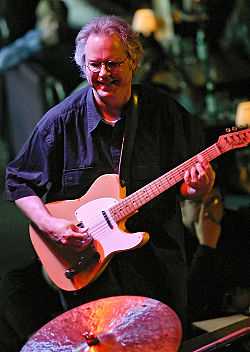 A man wearing glasses, playing a guitar and standing behind a cymbal.