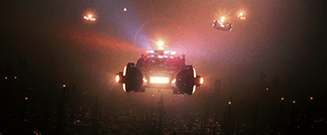 A screenshot from the film shows a line of police vehicles with flashing lights flying high above a smog-covered cityscape. Below them several small pinpoints of light from aircraft-avoidance lights on the tops of towers are all that can be seen of the city