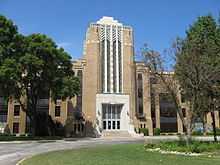 brown and white multi-story building, Art Deco style, with stone statues on each side of the steps, a circular drive in front, and green trees on the sides and center island of the drive