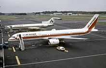 Side view of twin-engine jet on tarmac, with attached airstairs and support vehicle, along with a trijet aircraft in the background.