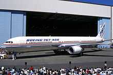 Side quarter view of twin-engine jetliner in front of hangar, with surrounding crowds.