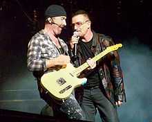 The Edge and Bono stand on a darkened stage, with lit-up smoke behind them. The Edge is strumming a guitar while Bono holds a microphone to his mouth.