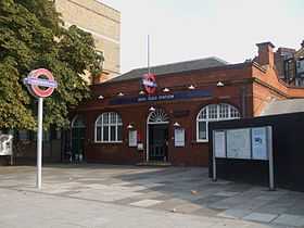A red-bricked building with a blue sign reading "BOW ROAD STATION" in white letters and a tree in the foreground all under a blue sky with white clouds