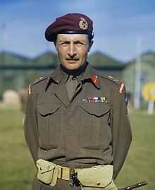 View from waist up of a man with short hair and moustache in battle dress with campaign ribbons. He is wearing a tie, airborne shoulder tabs, a maroon beret with a general's badge on it, and major-general's rank badges.