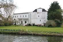 Colour photograph of a large white building with a castellated roof surrounded by trees situated in front of a river