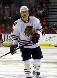 Hockey player in white uniform. He stands in a relaxed stance, one foot slightly off the ground.