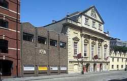 An imposing eighteenth-century building with three entrance archways, large first-floor windows and an ornate peaked gable end above. On the left, a twentieth-century grey brick building with a gilded crest; on the right a cream-coloured building with four pitched roofs. In front, a cobbled street.