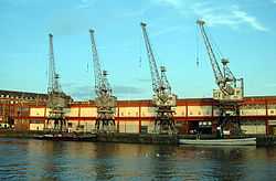 A long two-storey building with 4 cranes in front on the quayside. Two tugboats are moored at the quay.