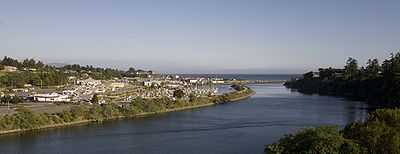 A wide, smooth river flows through the center. Its right bank is steep and forested, while its left bank is developed with many buildings and roads, and a marina filled with sailboats. Jetties and the Pacific Ocean are visible in the background, along with a hazy gray sky.