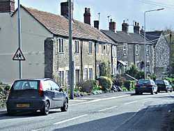 Street scene showing gray stone houses on the left of a road with a few cars.