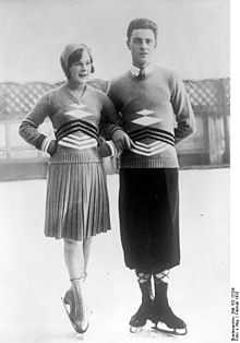 A woman and a man in figure skating blades stand on an outdoor ice rink posing for a shot. On the left, the woman has both hands in her waist, while the man has his right arm around her left arm.