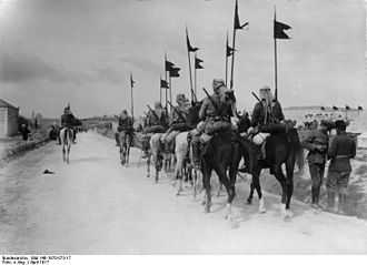 Black and white photo of mounted soldiers with middle eastern headwraps, carrying rifles, walking down a road away from the camera