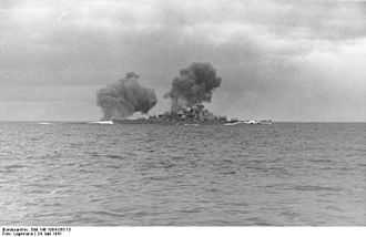 Black-and-white photograph of a warship firing the guns. Two columns of dark smoke drift from the ship