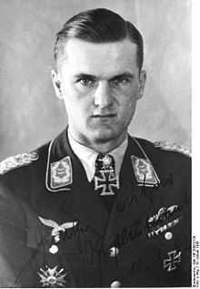 The head and shoulders of a young man, shown in semi-profile. He wears a military uniform with an Iron Cross displayed at the front of his shirt collar. His hair isshort and combed to his right, his nose is short, and his facial expression is determined; looking into the camera.