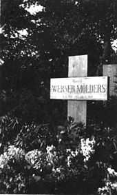 Black-and-white photograph of a wooden cross on a grave, bearing the inscription "Oberst Werner Mölders, 18. 3. 1913 – 22. 11. 1944." The name Werner Mölders is in large letters. Trees are seen in the background; the area in front of the cross is covered with low-growing plants bearing flowers.