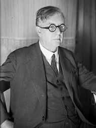 A middle-aged man in a suit, with slightly-unkempt, parted hair and small circular glasses
