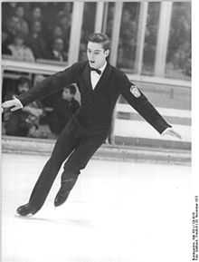 A young male figure skater is performing in an ice rink with a crowd in the background stands. He wears a formal black suit with white shirt and black bow tie, and his short hair is well combed.