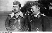 Black-and-white photograph showing half-length view of two uniformed men outdoors, standing next to each other. The young man on the left is wearing a field cap and a pilot's leather jacket with fur collar, with an Iron Cross displayed at the front of his shirt collar. The middle-aged man on the right is shown in three-quarter profile; he is smiling and wears a jacket with military decorations.