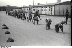 A line of half-naked prisoners performing "leap frog", under supervision of one of the Kapos. In the background the main gate to Mauthausen as well as two wooden barracks are visible.