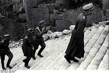 Four men dressed in SS uniforms climb the stone-cut stairs of death, one of them is smoking a cigarette.