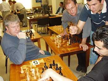 A picture of four men looking over a chessboard and gesticulating. In the background are other chess boards on tables, set up differently.